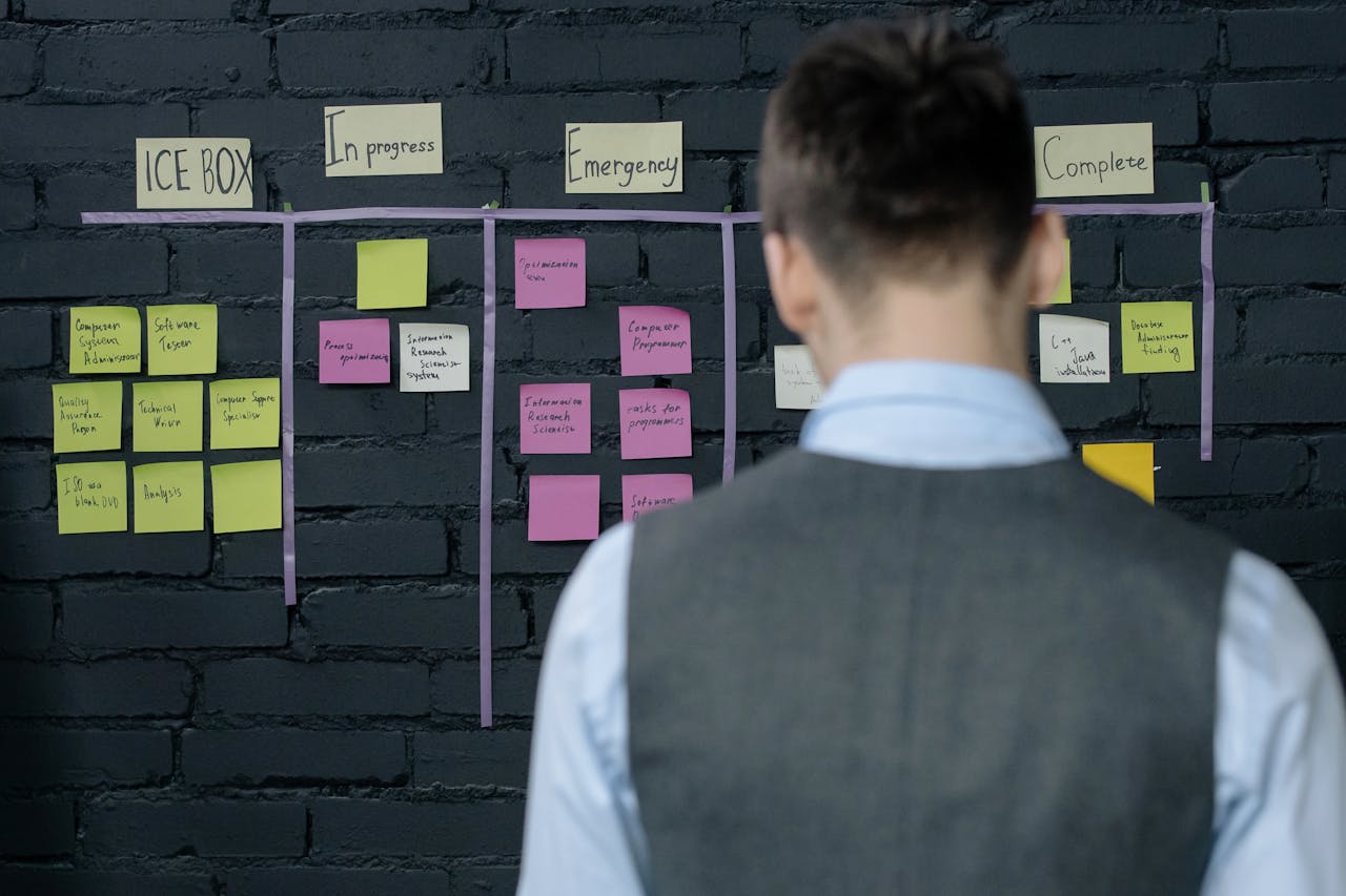 A person in a vest stands in front of a project management board with sticky notes on a dark brick wall.